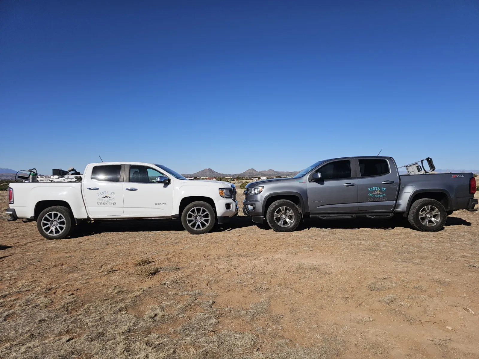 Two Santa Fe Pest Control pickup trucks, lettered with the company name and phone number, parked on open high-desert ground under a clear blue sky